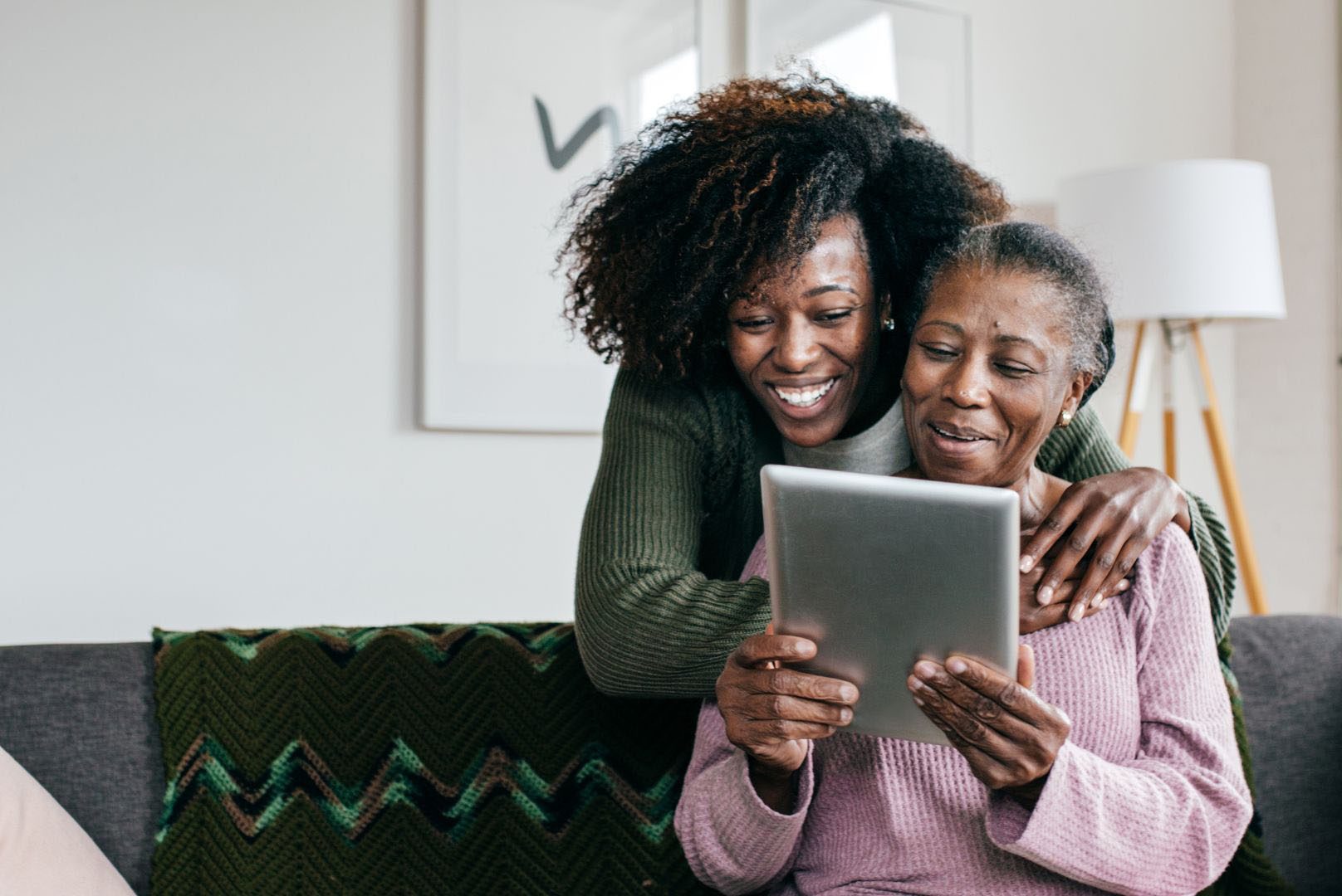 Young woman and her elderly mother sitting on a couch, smiling while looking at a tablet together